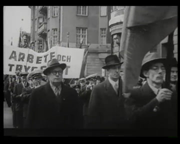 Schwarz-Weiß-Aufnahme aus der Schwedischen Wochenschau von der Ersten Mai-Kundgebung im Jahr 1944. Im Hintergrund sieht man eine Marschkapelle und ein Transparent mit der Aufschrift: „Arbete och trygghet“.,