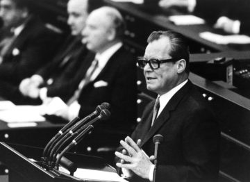 Schwarz-Weiß-Aufnahme von Willy Brandt aus dem Jahre 1969 vor einem Rednerpult im Bonner Bundestag. Links neben ihm ist unscharf auf der Regierungsbank Walter Scheel zu sehen.